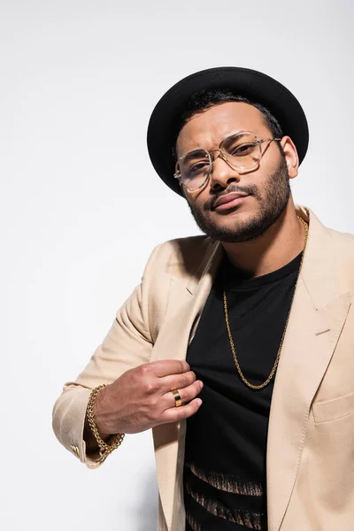 Elegante artista indio de hip hop en sombrero fedora y gafas graduadas ajustando chaqueta beige aislado en gris - foto de stock