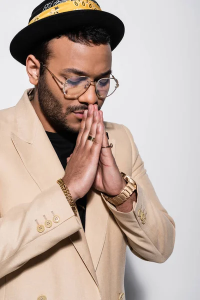 Stylish indian hip hop singer in fedora hat and eyeglasses praying isolated on grey - foto de stock