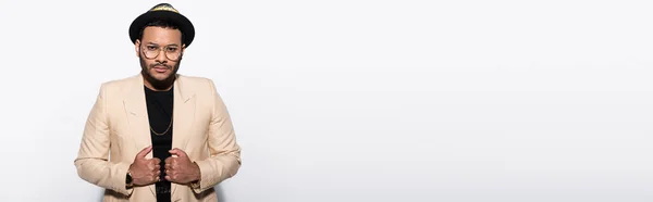 Indian hip hop singer in fedora hat and eyeglasses adjusting blazer isolated on grey, banner — Stock Photo