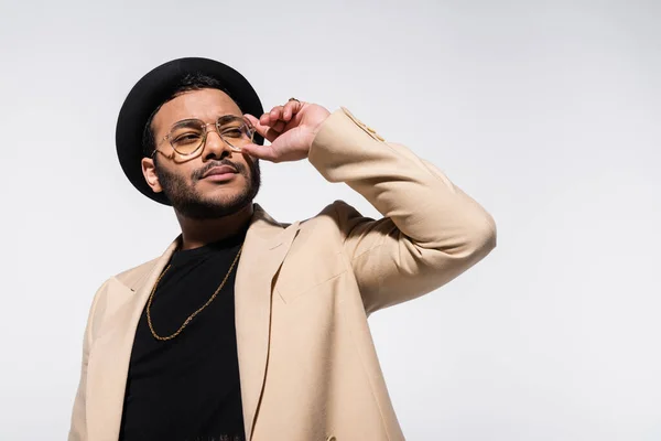 Elegante artista indio de hip hop en sombrero fedora ajustando gafas aisladas en gris - foto de stock