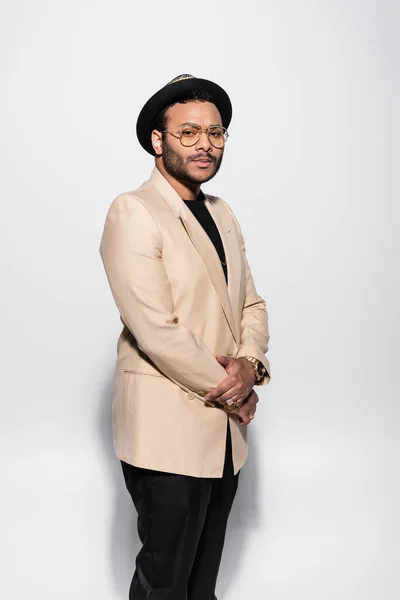 Stylish indian hip hop singer in fedora hat and eyeglasses standing isolated on grey — Photo de stock