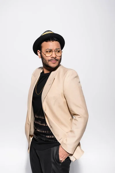 Stylish indian hip hop performer in fedora hat and eyeglasses posing with hand in pocket isolated on grey — Fotografia de Stock