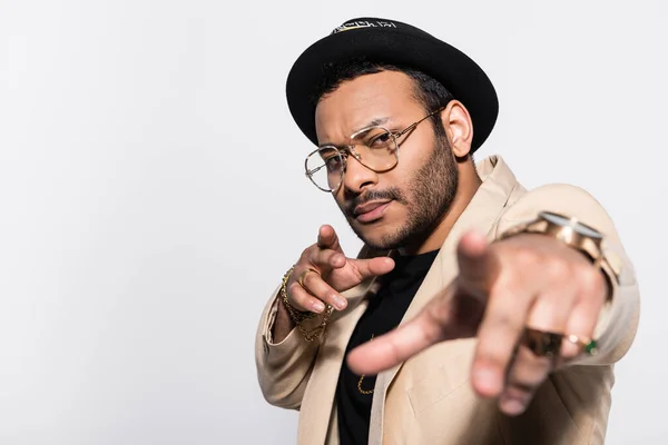 Elegante artista indio de hip hop en sombrero fedora y gafas apuntando a la cámara aislada en gris - foto de stock