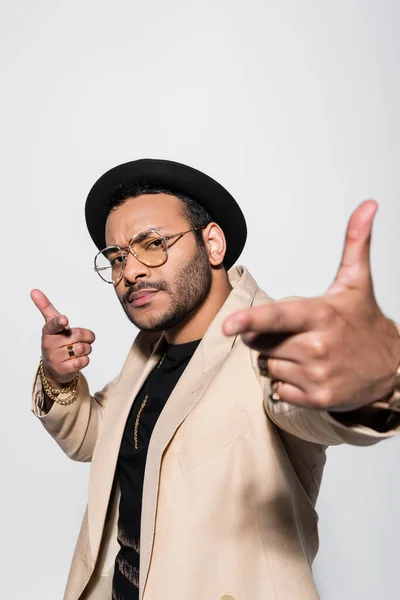 Stylish indian hip hop singer in hat and eyeglasses pointing at camera isolated on grey — Foto stock