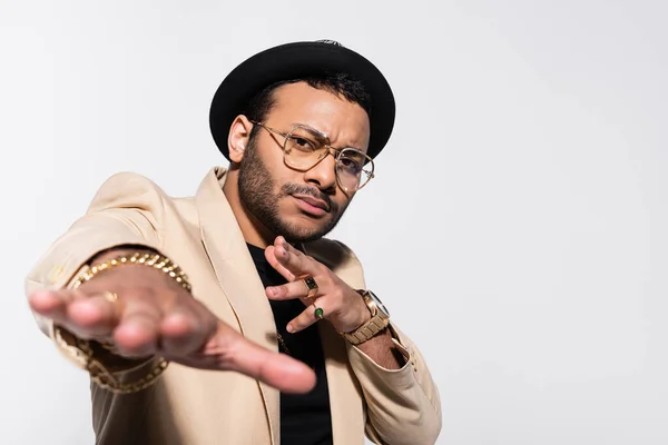 Stylish indian hip hop singer in hat and eyeglasses gesturing and looking at camera isolated on grey — Stock Photo