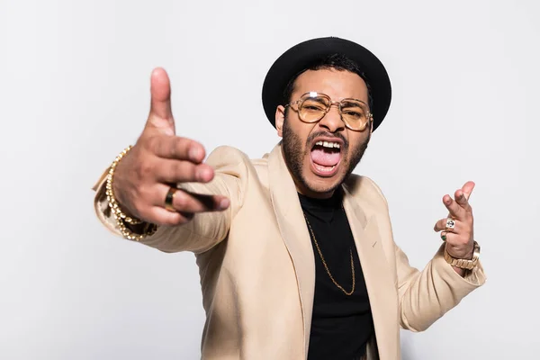 Emotional indian hip hop performer in fedora hat and eyeglasses screaming while gesturing isolated on grey — Photo de stock