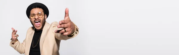 Emotional indian hip hop performer in fedora hat and eyeglasses screaming while gesturing isolated on grey, banner — Stock Photo