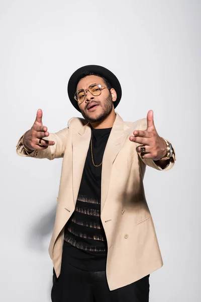 Cool cantante indio de hip hop en sombrero y gafas gestos y mirando a la cámara en gris - foto de stock