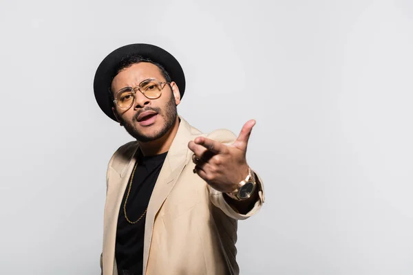 Indian hip hop performer in fedora hat and eyeglasses pointing with finger isolated on grey — Stockfoto