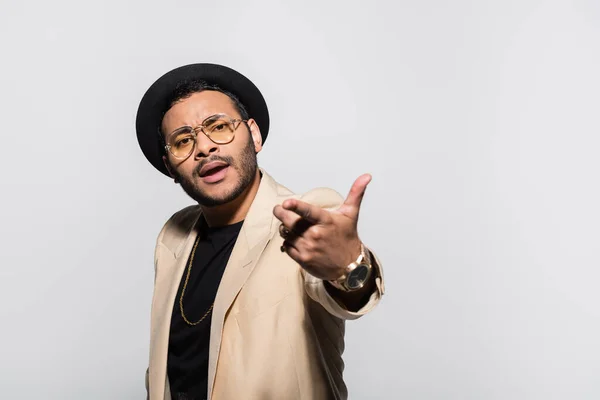 Indian hip hop performer in fedora hat and eyeglasses pointing with finger isolated on grey — Stock Photo