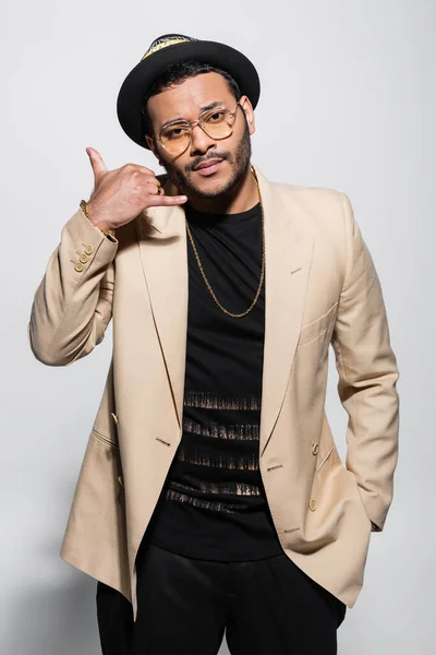 Bearded indian hip hop performer in fedora hat and eyeglasses showing call me gesture on grey — Stock Photo