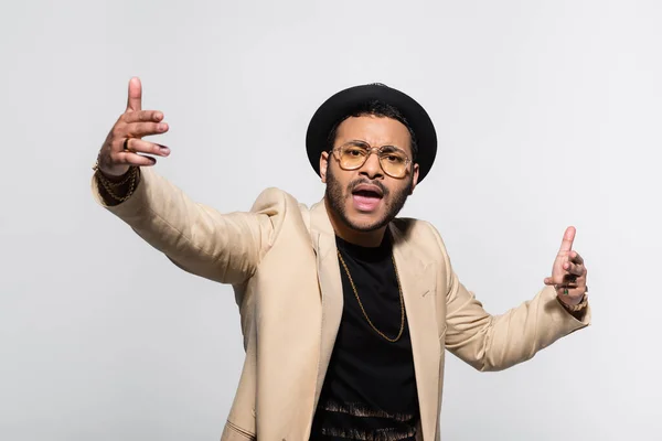 Eastern hip hop performer in fedora hat and eyeglasses singing while gesturing isolated on grey — Stockfoto