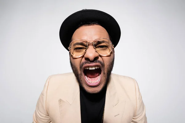 Emotional indian hip hop performer in fedora hat and eyeglasses screaming isolated on grey - foto de stock