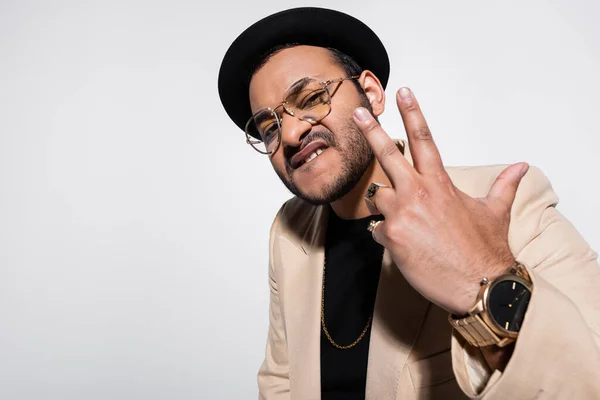 Eastern hip hop performer in fedora hat and eyeglasses biting lip and gesturing isolated on grey — Stock Photo