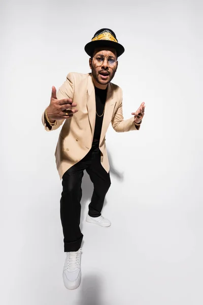 Full length of cool eastern hip hop performer in fedora hat and eyeglasses gesturing on grey — Stock Photo