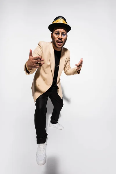 Full length of cool eastern hip hop performer in fedora hat and eyeglasses gesturing on grey — Foto stock