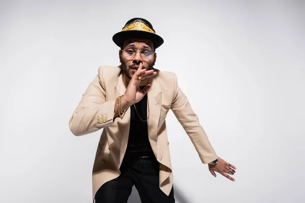 Artista de hip hop oriental con sombrero de sombrero de sombrero de sombrero y gafas que muestran el signo de silencio en gris - foto de stock