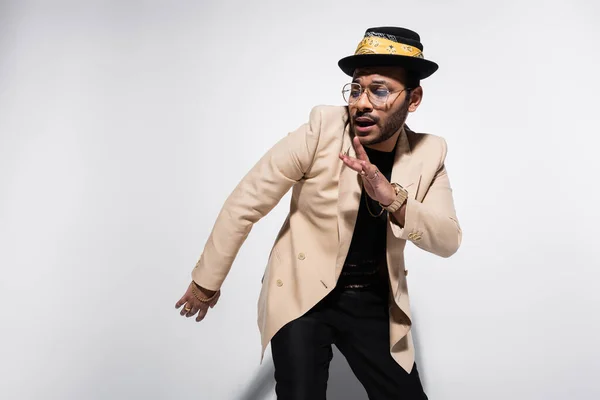 Восточный хип-хоп исполнитель в шляпе и очках, смотрящий в сторону и жестикулирующий на серый — стоковое фото