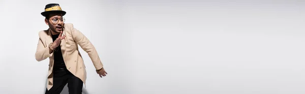 Artista de hip hop oriental en sombrero fedora y anteojos mirando hacia otro lado y haciendo gestos en gris, bandera - foto de stock