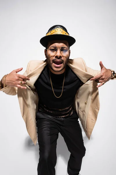 Eastern hip hop performer in fedora hat and eyeglasses gesturing while singing on grey — Foto stock