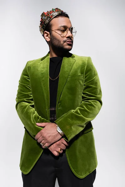 Восточный хип-хоп исполнитель в зеленом бархатном пиджаке и короне, смотрящий вдаль изолированный на сером — стоковое фото