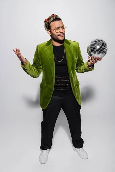 Смущенный восточный хип-хоп исполнитель в зеленом бархатном пиджаке и короне, держащий диско-мяч на сером — стоковое фото