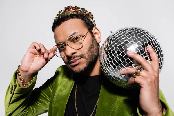 Eastern hip hop performer in green velvet blazer and crown holding disco ball while adjusting eyeglasses isolated on grey — Stock Photo