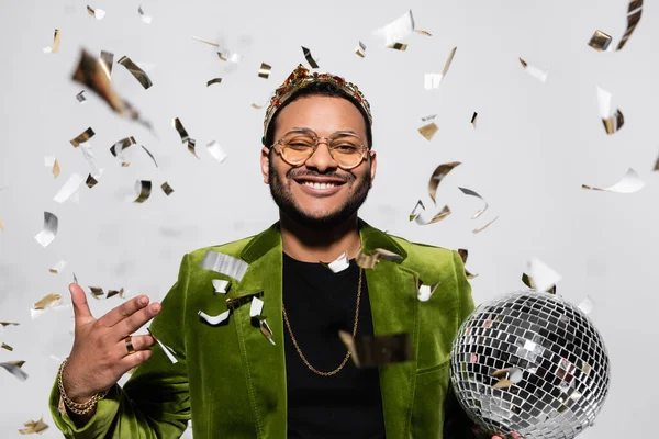 Artista de hip hop indio feliz en chaqueta de terciopelo verde y corona sosteniendo bola disco cerca de caer confeti en gris - foto de stock