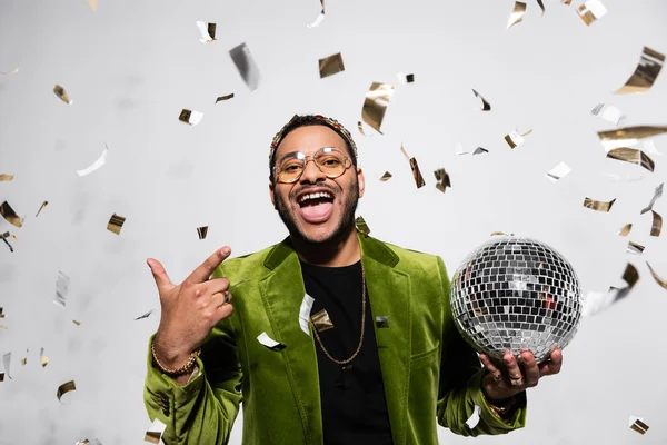Amazed indian hip hop performer in green velvet blazer and crown holding disco ball near falling confetti on grey — Stock Photo
