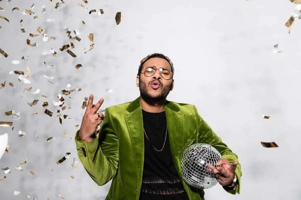 Indian hip hop performer in crown showing peace near falling confetti on grey — Stock Photo