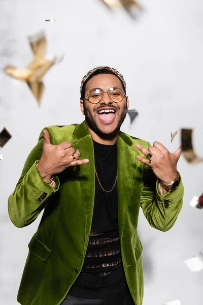 Excited indian hip hop performer in green velvet blazer and crown gesturing near falling confetti on grey — Photo de stock