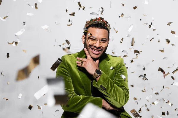 Alegre indiana hip hop performer em blazer de veludo verde e coroa perto de cair confetti em cinza — Fotografia de Stock