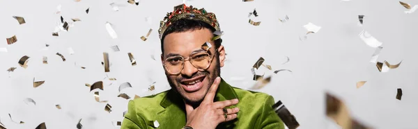 Cheerful indian hip hop performer in green velvet blazer and crown near falling confetti on grey, banner - foto de stock