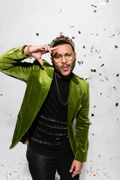 Artista indio de hip hop en chaqueta de terciopelo verde y la corona haciendo pucheros labios cerca de caer confeti en gris - foto de stock