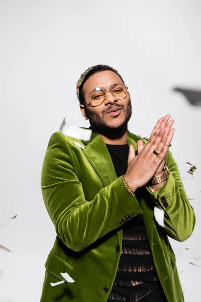 Indian hip hop performer in green blazer and crown looking at camera near falling confetti on grey — Stock Photo