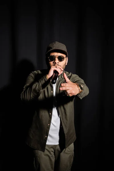 Eastern hip hop performer in sunglasses singing in microphone and pointing at camera on black - foto de stock