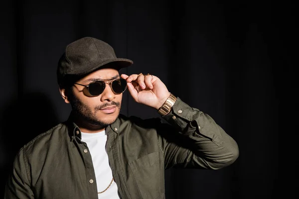 Eastern hip hop performer in cap adjusting sunglasses on black — Stockfoto