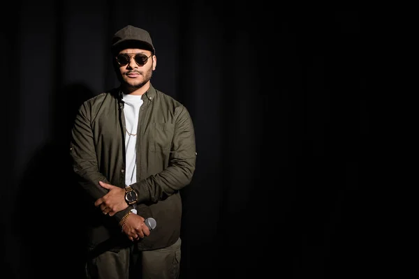 Восточный хип-хоп исполнитель в солнцезащитных очках с микрофоном на черном — стоковое фото