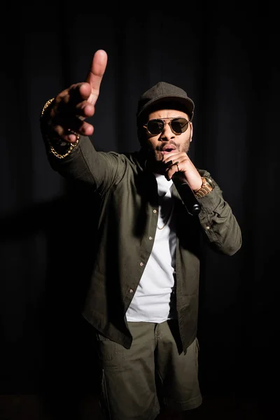 Східний хіп-хоп виконавець в сонцезахисних окулярах жестикулює під час співу в мікрофоні на чорному — стокове фото