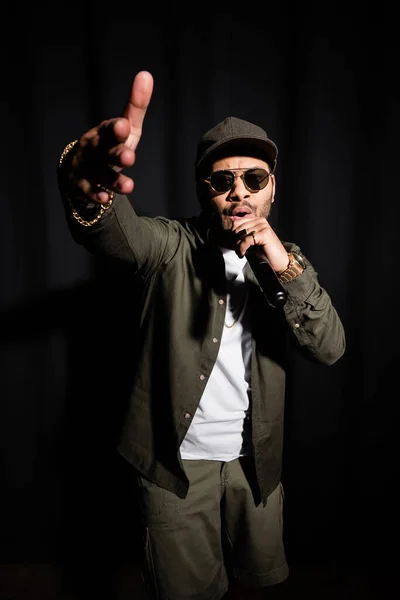 Eastern hip hop performer in sunglasses gesturing while singing in microphone on black — Fotografia de Stock