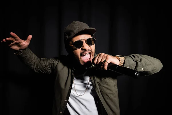 Cantante de hip hop oriental en gafas de sol cantando en micrófono en negro - foto de stock