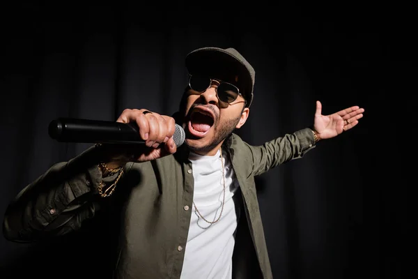 Emotional eastern hip hop performer in sunglasses singing in microphone on black - foto de stock