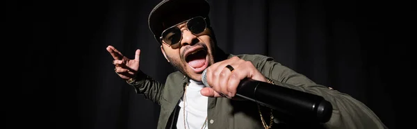 Indian hip hop performer in sunglasses and cap singing in microphone on black, banner — Stockfoto