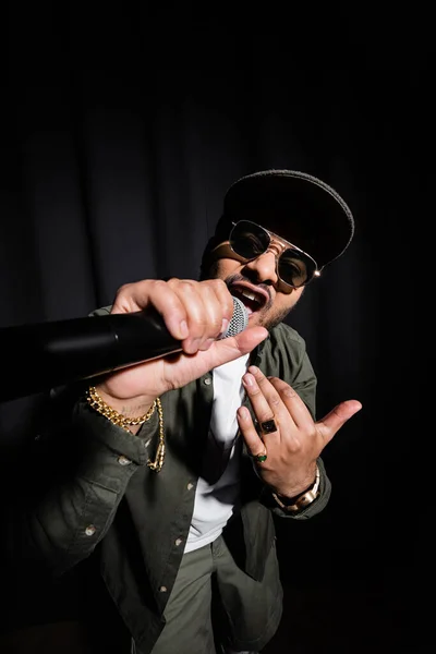 Индийский хип-хоп исполнитель в солнцезащитных очках поющий в микрофоне на черном — стоковое фото