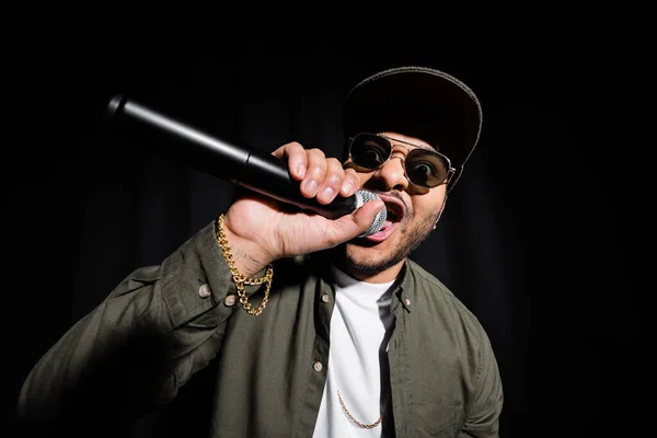 Emotional middle east hip hop singer in sunglasses and cap singing in microphone and gesturing isolated on black — Foto stock