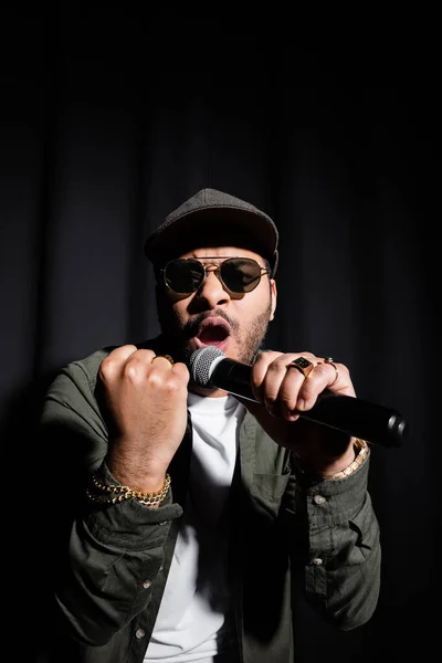 Emotional middle east hip hop performer with clenched fist singing in microphone on black - foto de stock