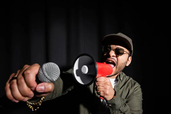 Эмоциональный ближневосточный хип-хоп исполнитель в солнечных очках и кепке поющий в микрофоне и громкоговоритель на черном — стоковое фото