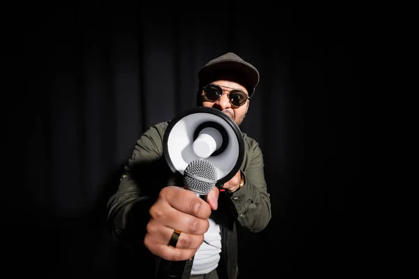 Ближневосточный хип-хоп исполнитель в солнцезащитных очках и кепке поющий в микрофоне и громкоговоритель на черном — стоковое фото