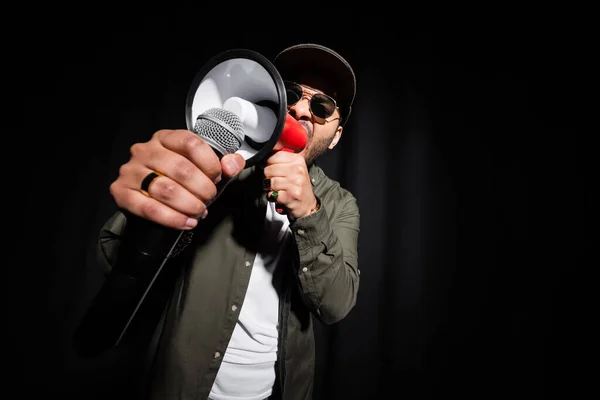 Cantante de hip hop de Oriente Medio en gafas de sol y gorra cantando en micrófono y altavoz en negro - foto de stock