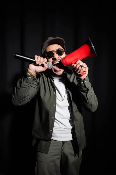 Emotional indian hip hop performer in sunglasses and cap screaming in microphone and loudspeaker on black - foto de stock